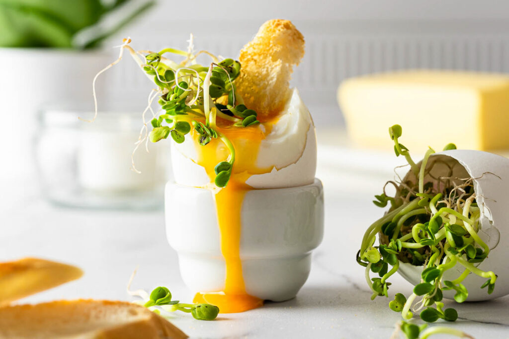 Et bilde av et egg i et egg holder, med den gyldne plommen rennende nedover holderen. Ved siden av ligger ristet brød og delikate spirer i et eggskall. Bilder er lys og luftig.