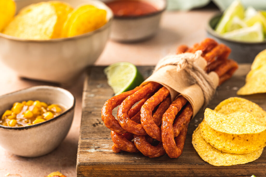 Et bilde av et assortert utvalg av snacks, inkludert snacks pølse, marinert mais, quacamole, taco chips og annet fingeremat, servert på et fat og i skål